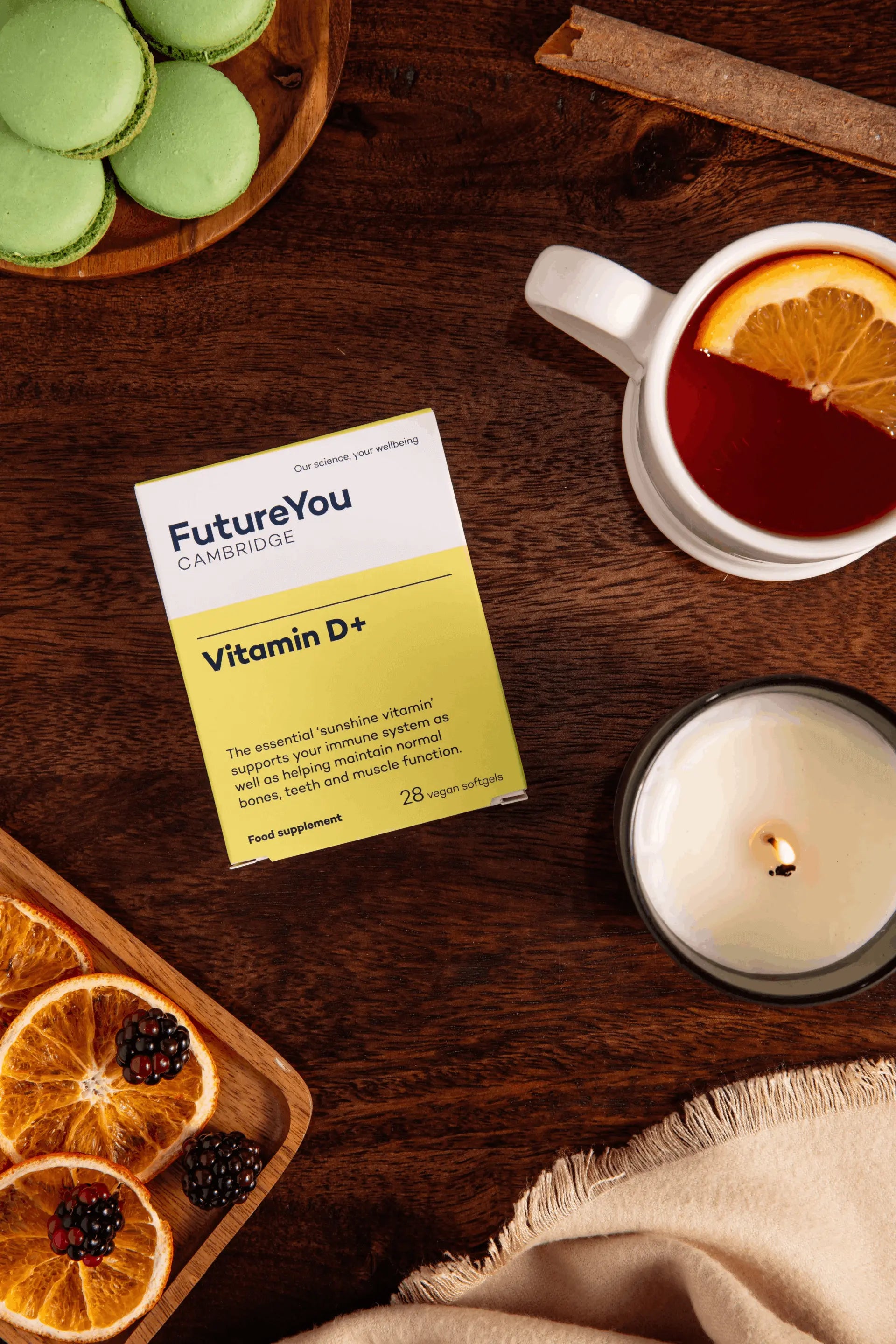 Vitamin D supplement package on a wooden table with tea, oranges, and a candle.